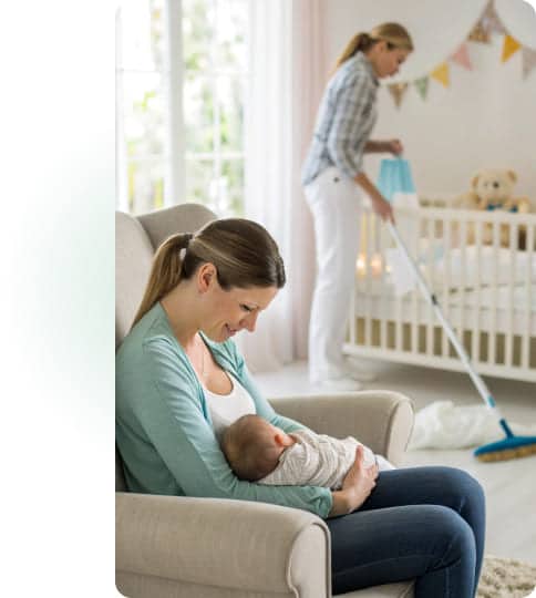 Eine Mutter stillt ihr Baby im Kinderzimmer, während im Hintergrund eine Reinigungskraft den Boden reinigt – Unterstützung im Alltag mit einem Neugeborenen.