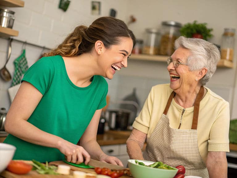 Eine Alltagsfee und eine Seniorin lachen gemeinsam bei der Zubereitung einer Mahlzeit.